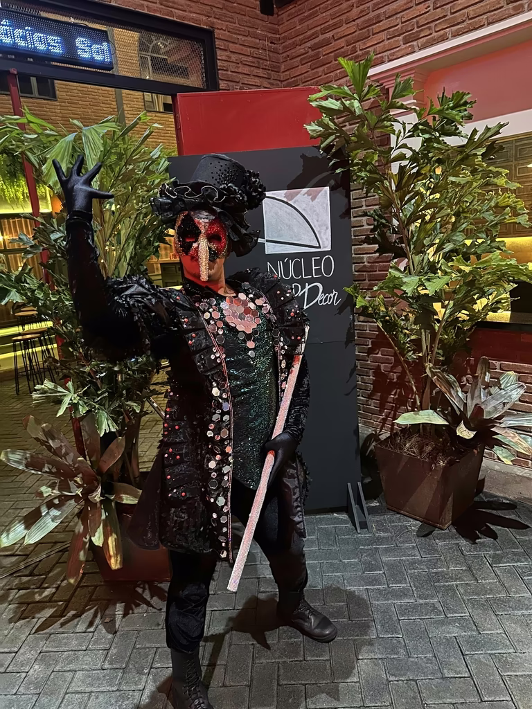 Performer mascarado em traje veneziano diante do painel do Núcleo Top Decor