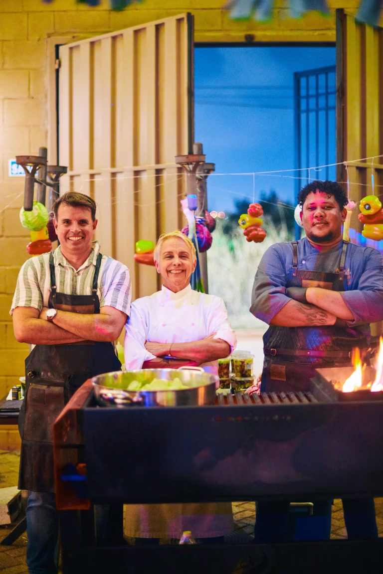Três chefs sorridentes de avental posam atrás da grelha com guirlandas de legumes